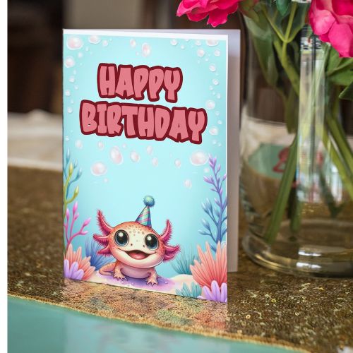 Birthday card with a cartoon axolotl and 'Happy Birthday' text, placed on a decorative surface with flowers.